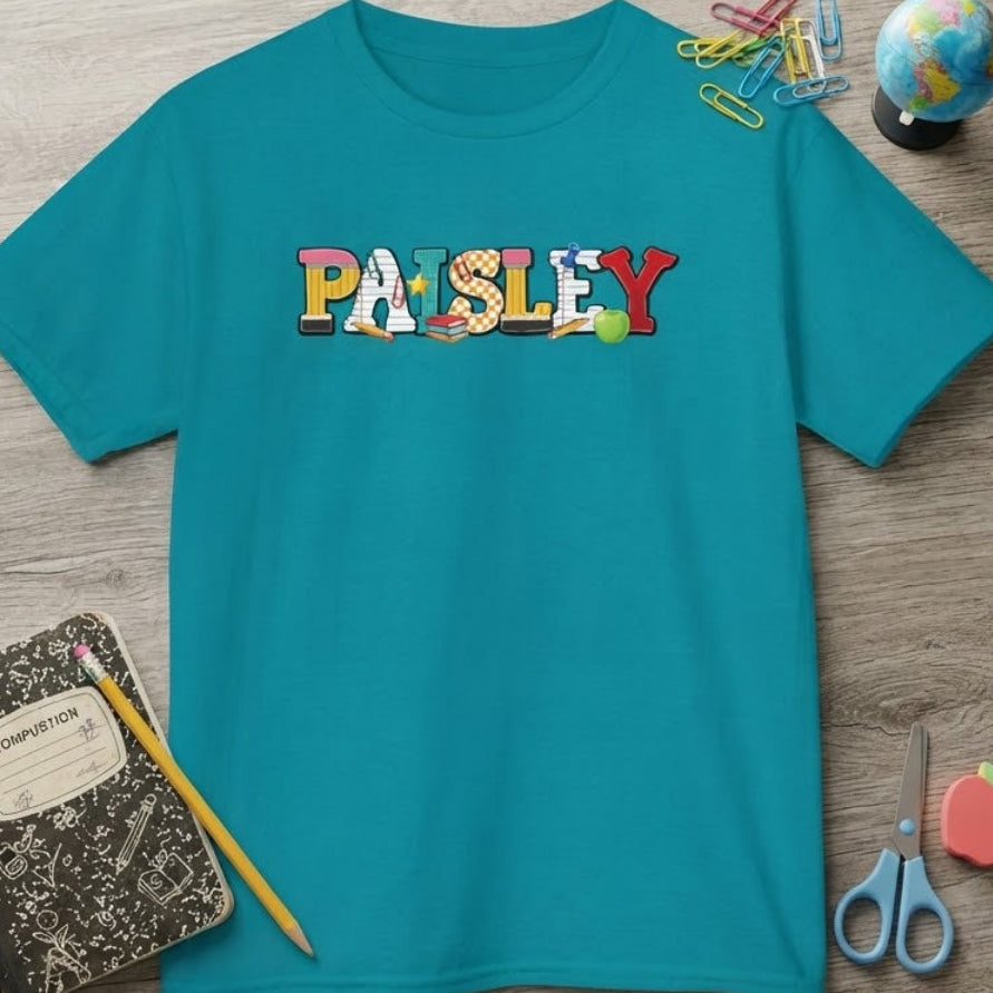 A turquoise short-sleeve T-shirt laid flat on a wood desk with school-themed props including a notebook, pencil, paperclips, a small globe, scissors, and a fruit eraser. The shirt has a colorful 'PAISLEY' graphic across the chest.