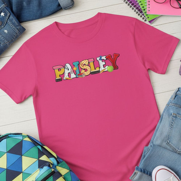 A bright pink short-sleeve t-shirt with the word 'PAISLEY' printed across the chest in colorful school-style letters. The shirt is styled in a flat lay on a white wood background with jeans, a backpack, and notebooks around it.