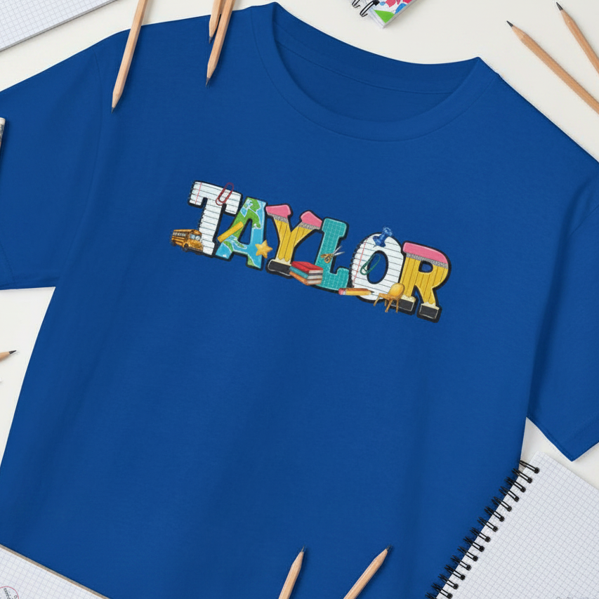 A royal blue short-sleeve t-shirt laid flat on a white desk with pencils and notebooks around it. The front has a colorful &