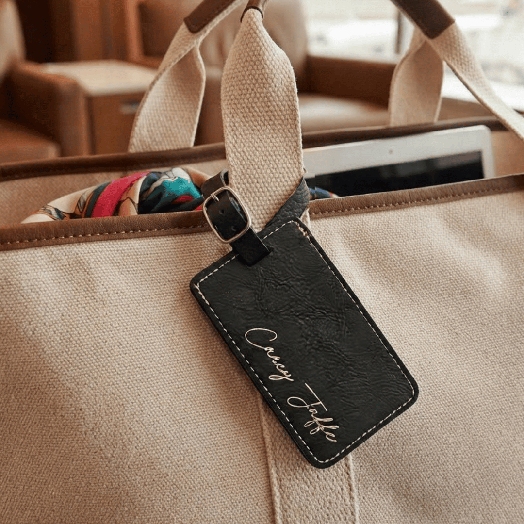 Beige bag with a black leather luggage tag attached, featuring cursive text name engraved in metallic gold.