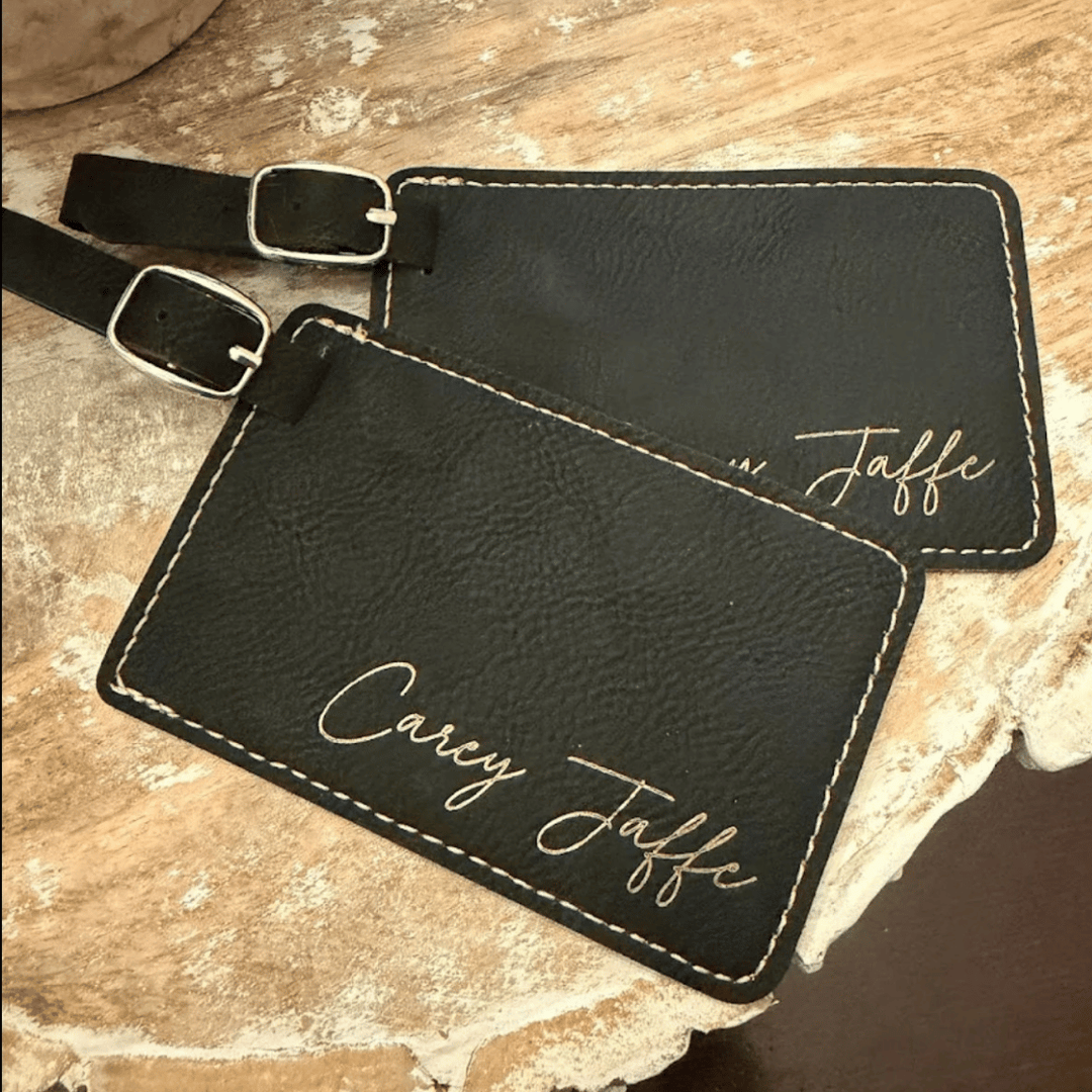 Two black leather luggage tags with gold text engraved in metallic gold, laying on a textured surface