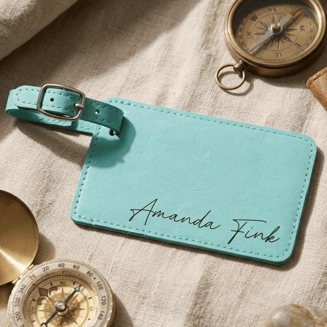 Teal luggage tag with 'Amanda Fink' engraved in black, surrounded by vintage compasses on a textured surface.