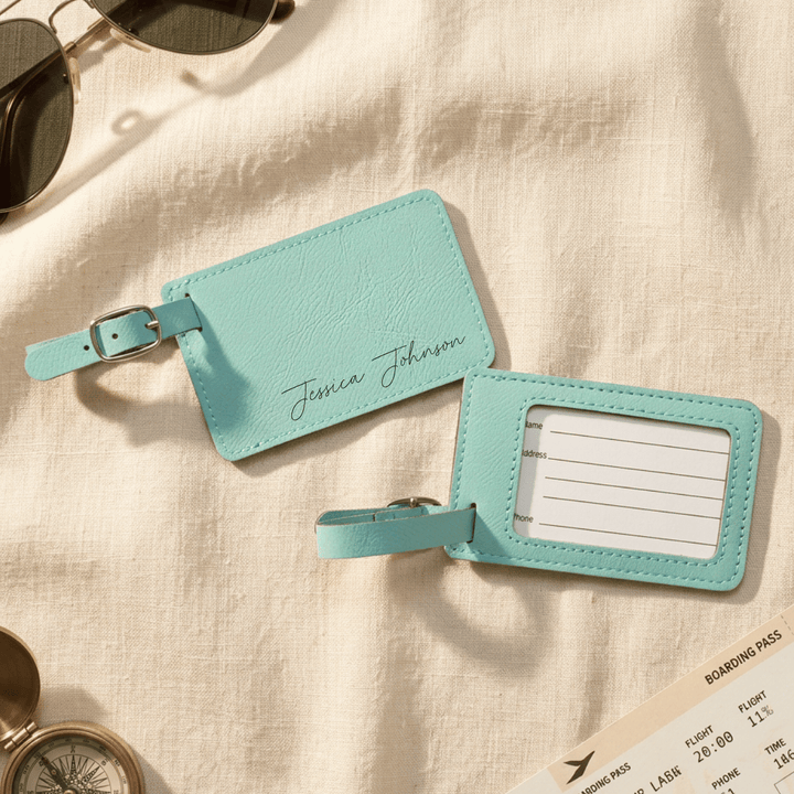 Front and back of teal luggage tags showing engraved name on  front and clear plastic back with personalization insert, laying on a beige fabric background