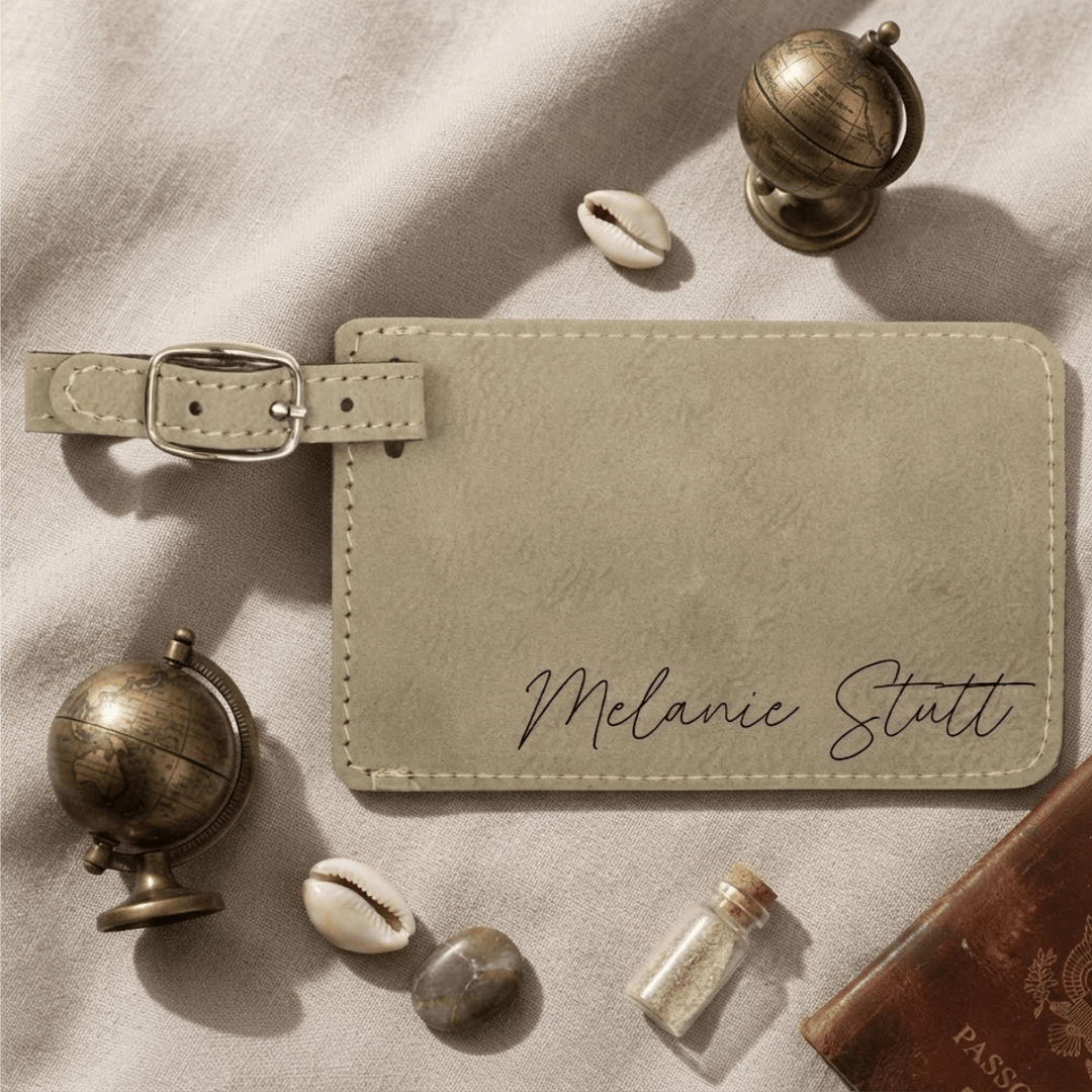 Light brown leather luggage tag with 'Melanie Stutt' engraving in black script, surrounded by decorative items on a textured surface.