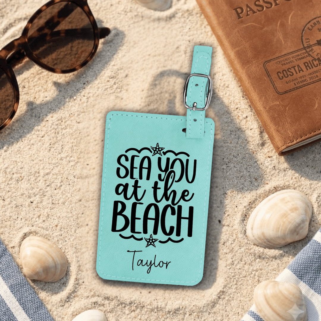Turquoise luggage tag with 'Sea you at the Beach' text on sand with sunglasses, passport, and shells.