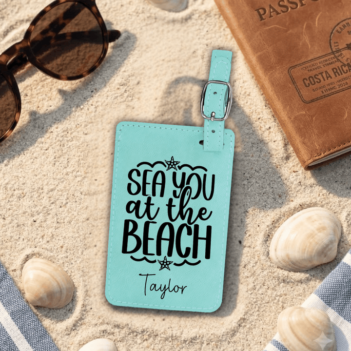 Turquoise luggage tag with 'Sea you at the Beach' text on sand with sunglasses, passport, and shells.