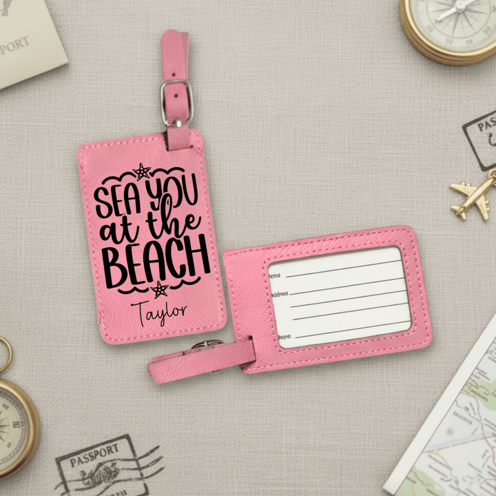 Pink luggage tag with 'Sea you at the Beach' text on a textured surface with travel-themed items.