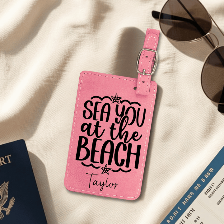 Pink luggage tag with 'Sea you at the Beach Taylor' text on a beige surface with sunglasses and passport.
