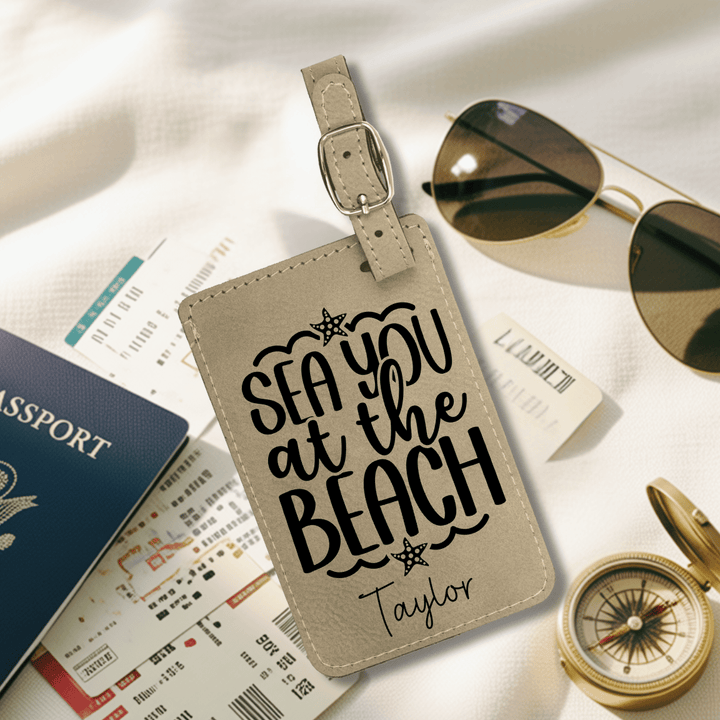 Beige luggage tag with 'Sea you at the Beach Taylor' text on a travel-themed background with passport, sunglasses, and compass.