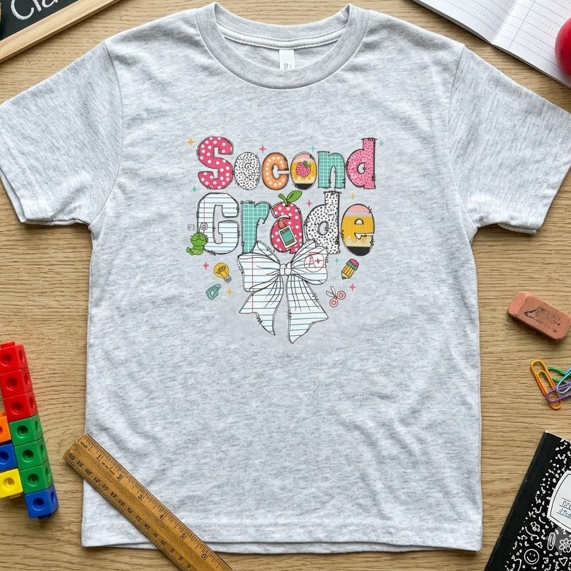 A light heather gray short-sleeve t-shirt with a colorful 'Second Grade' bow graphic, styled in a flat-lay on a wooden desk with school supplies including a chalkboard, notebooks, pencils, and a ruler.