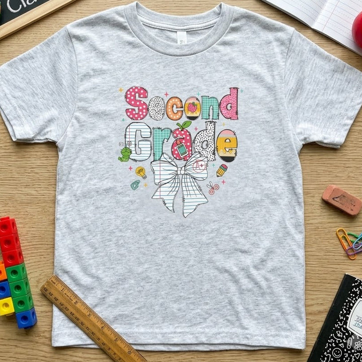 A light heather gray short-sleeve t-shirt with a colorful 'Second Grade' bow graphic, styled in a flat-lay on a wooden desk with school supplies including a chalkboard, notebooks, pencils, and a ruler.