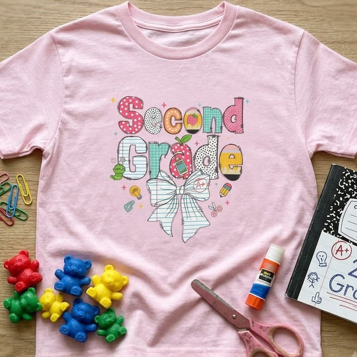 A light pink short-sleeve t-shirt with a colorful 'Second Grade' bow graphic, styled in a flat-lay on a light wood background with school supplies including a composition book, scissors, paper clips, and candy.