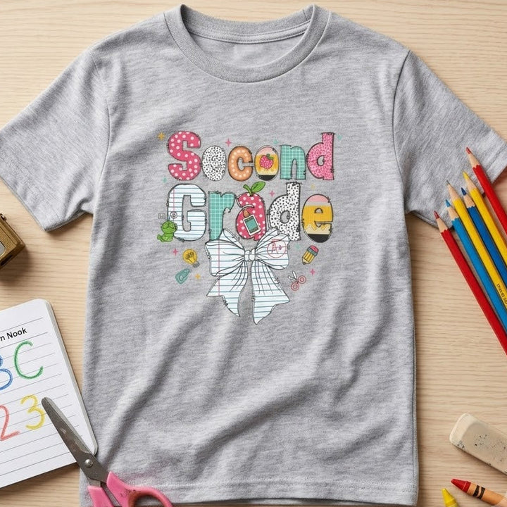A heather gray short-sleeve t-shirt with a colorful 'Second Grade' bow graphic, styled in a flat-lay on a light wood background with school supplies including pencils, an apple, scissors, a composition book, and an eraser.