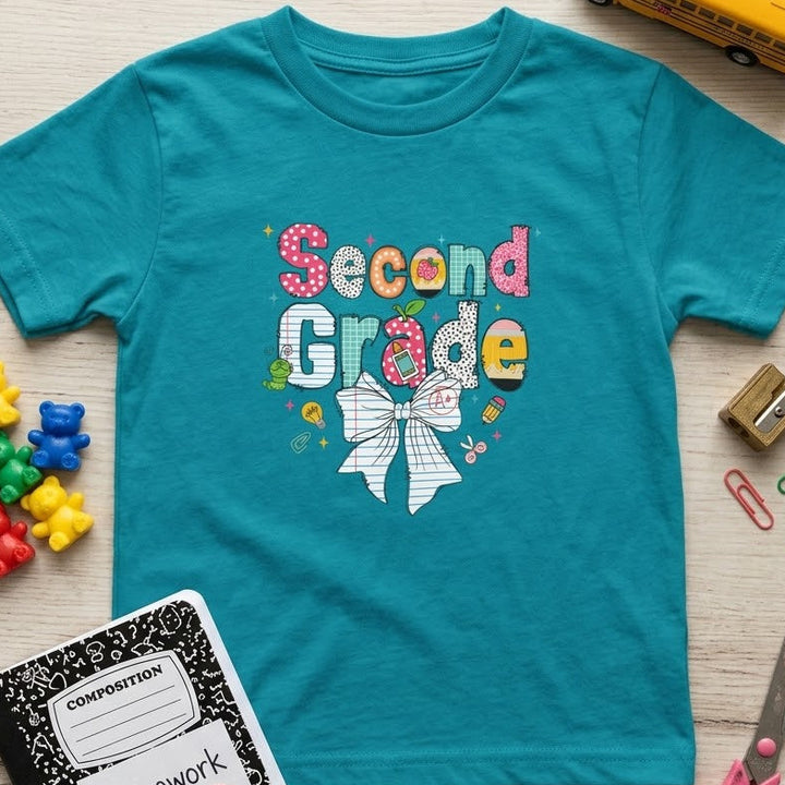 A teal short-sleeve t-shirt with a colorful 'Second Grade' bow graphic, laid flat on a light wood background surrounded by school-themed props including a yellow school bus, notebook, scissors, paper clips, and small toy bears.