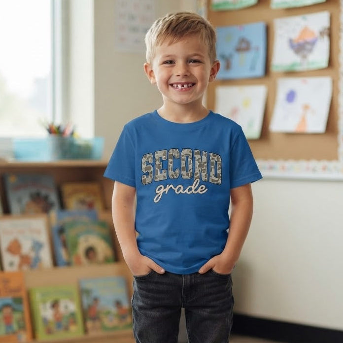 A young boy stands in a bright classroom wearing a blue graphic t-shirt that reads 'SECOND grade' in camo block letters and tan script, paired with dark jeans and sneakers.
