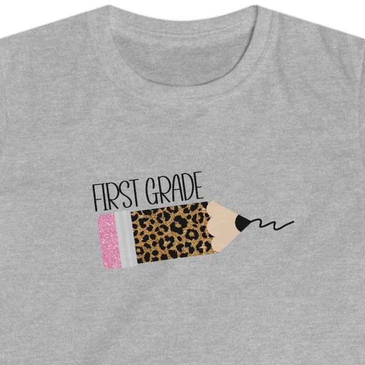 A heather gray short-sleeve t-shirt with a graphic on the chest depicting a pencil with a leopard print body, a pink eraser, and the text 'FIRST GRADE' alongside a scribble, shown on a plain white background.