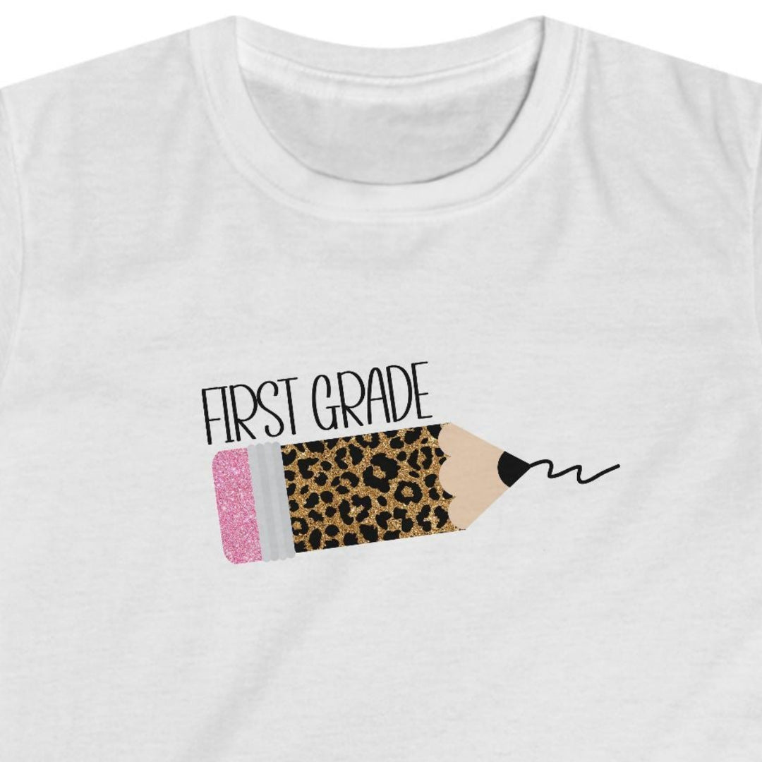 A white short-sleeve crewneck t-shirt featuring a graphic of a pencil with a leopard print body, a pink eraser, and the text 'FIRST GRADE' printed above it.