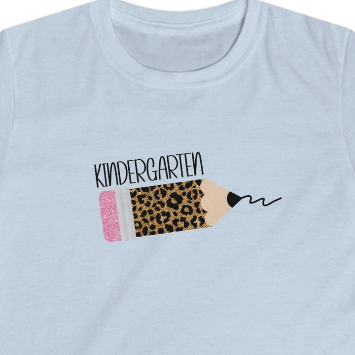 A light blue short-sleeve kids' t-shirt with a leopard pencil graphic slanted on the chest featuring black text above it that says 'kindergarten', displayed on a clean white background.
