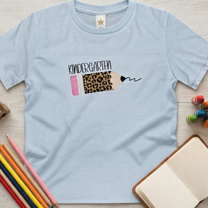 A light blue short-sleeve t-shirt laid flat on a light wood surface, featuring a sparkly leopard print pencil graphic with 'kindergarten' black text on top of pencil. The shirt is styled with school supplies including colored pencils, a sharpening stone, paintbrushes, and a small notebook.