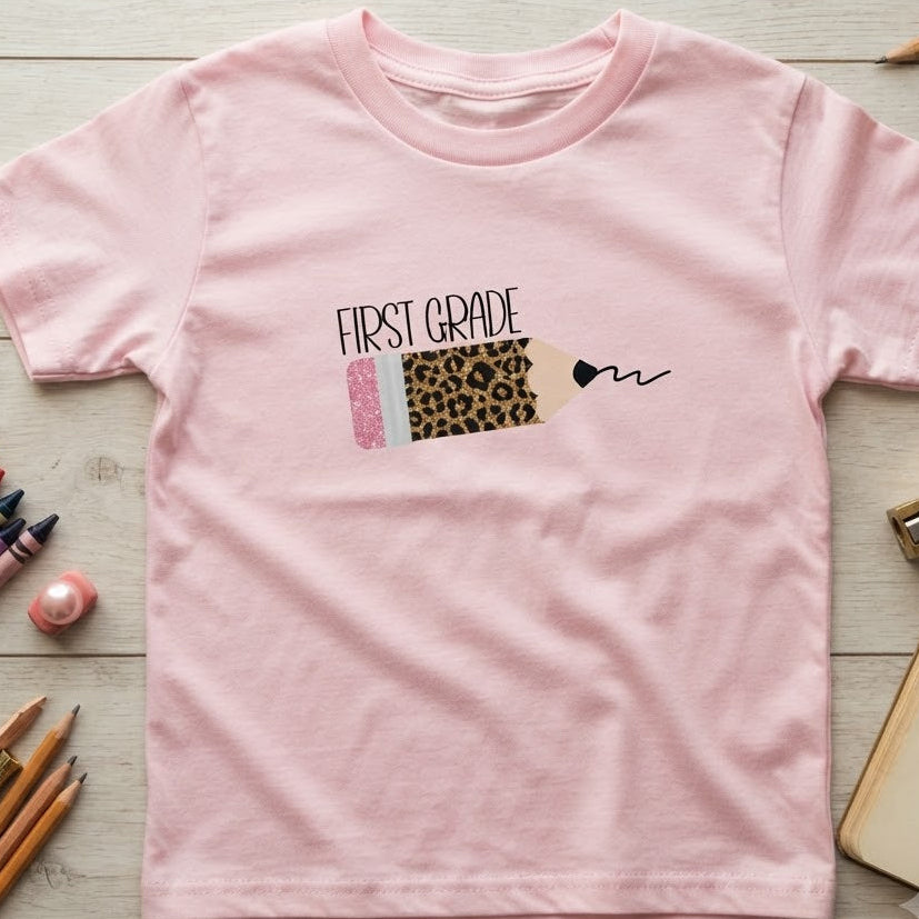 A light pink short-sleeve t-shirt for kids, featuring a sparkly leopard pencil graphic and black text on top of pencil that reads 'first grade'.  The t-shirt is laid flat on a light wood background surrounded by school supplies like colored pencils, a notebook, and a eraser. 