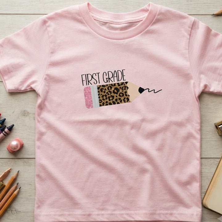 A light pink short-sleeve t-shirt for kids, featuring a sparkly leopard pencil graphic and black text on top of pencil that reads 'first grade'.  The t-shirt is laid flat on a light wood background surrounded by school supplies like colored pencils, a notebook, and a eraser. 