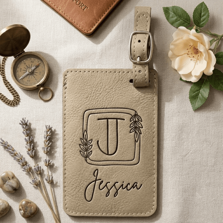 Beige leather luggage tag with engraved initials and name, surrounded by decorative items on a light surface.