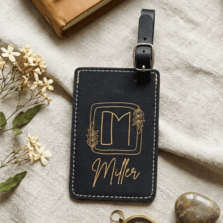 Black leather luggage tag with gold lettering on a textured surface with flowers and a book.