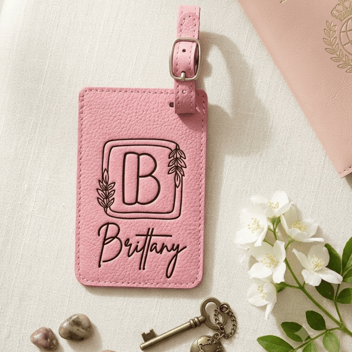 Pink leather luggage tag with 'B' and 'Brittany' engraved text, placed on a light surface with flowers and keys.