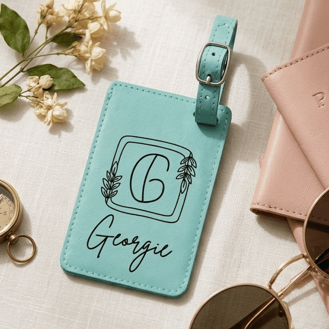 Turquoise luggage tag with 'Georgie' and a decorative 'G' engraved in black laying on a light surface with flowers and a pink wallet.