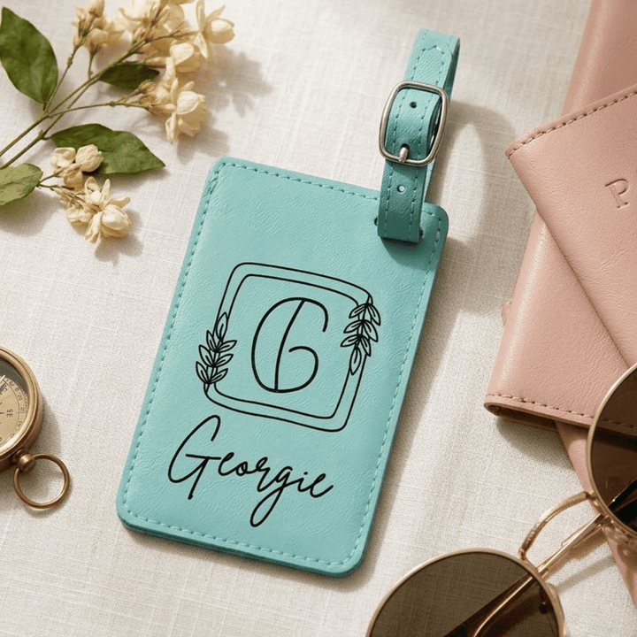 Turquoise luggage tag with 'Georgie' and a decorative 'G' engraved in black laying on a light surface with flowers and a pink wallet.