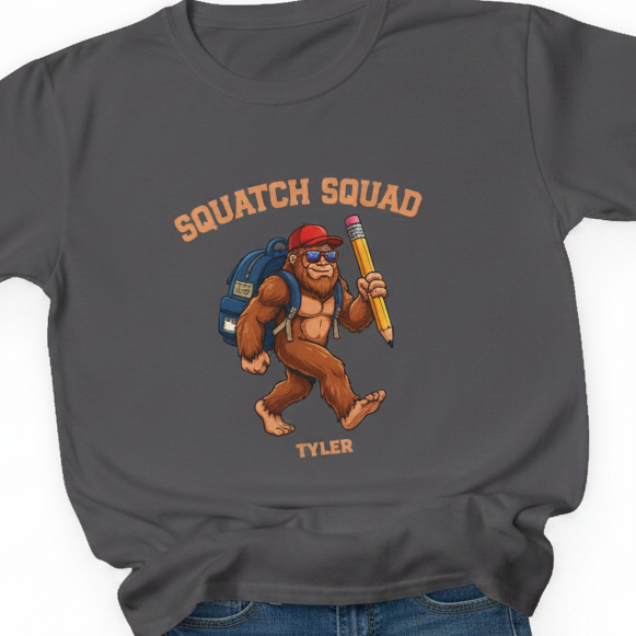 A dark charcoal gray crewneck t-shirt featuring a cartoon graphic of a brown furry Bigfoot-like character wearing a red cap and blue backpack, holding a giant pencil. The text 'SQUATCH SQUAD' is printed above in tan, and 'TYLER' is printed below.