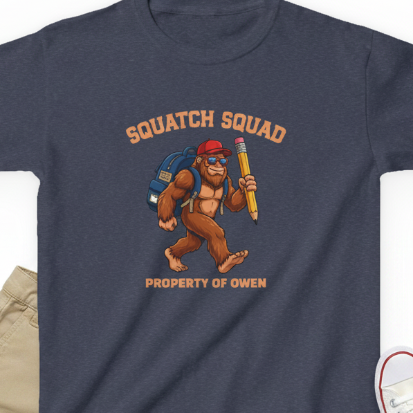 A navy blue heather graphic t-shirt featuring a cartoon Bigfoot wearing a red cap and sunglasses, holding a yellow pencil and carrying a blue backpack. The text 'SQUATCH SQUAD' is printed above the character, and 'PROPERTY OF OWEN' is printed below.