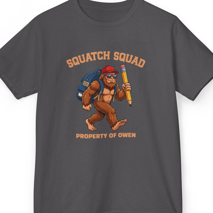 Charcoal t-shirt with 'Squatch Squad' collegiate text in an arc on top of a bigfoot cartoon character wearing a blue backpack, red hat, and holding a giant pencil. Bottom line of text is personalizable. This one says 'property of owen'
