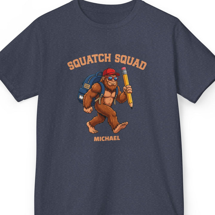 Heather navy t-shirt with 'Squatch Squad' collegiate text in an arc on top of a bigfoot cartoon character wearing a blue backpack, red hat, and holding a giant pencil. Bottom line of text is personalizable. This one says 'Michael'