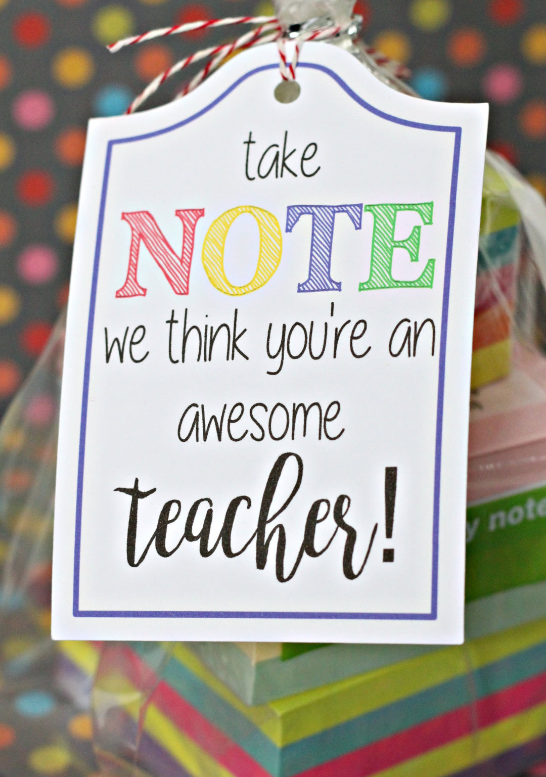 A white gift tag with the phrase 'take NOTE we think you're an awesome teacher!' in black and colorful lettering, hanging from a red-and-white string. The tag is shown in front of a blurred background with colorful confetti and stacked gift items.