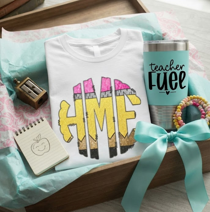 A styled gift set in a wooden tray featuring a white graphic t-shirt with a pencil-monogram design, a teal insulated tumbler with 'teacher Fuel' text, a notebook, a pencil sharpener, and a pastel hair tie, arranged on a light wood surface near a window.