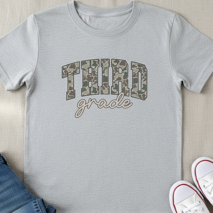 A light gray heather crewneck t-shirt with 'THIRD grade' printed on the front in camouflage block letters and cursive script. The shirt is styled in a flat lay with folded blue jeans and a pair of white low-top sneakers on a beige fabric background.