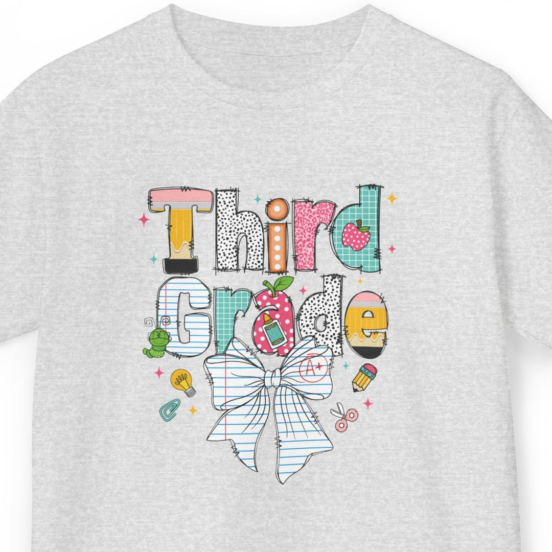 A light ash gray crewneck t-shirt featuring a colorful graphic design on the front. The graphic spells out 'Third Grade' in playful, hand-drawn lettering filled with school-themed illustrations like a pencil, apple, ruler, and a large striped bow.