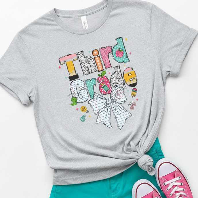 A light ash gray heather graphic T-shirt with a colorful 'Third Grade' bow design, styled in a flat-lay with teal shorts and pink sneakers on a white background.