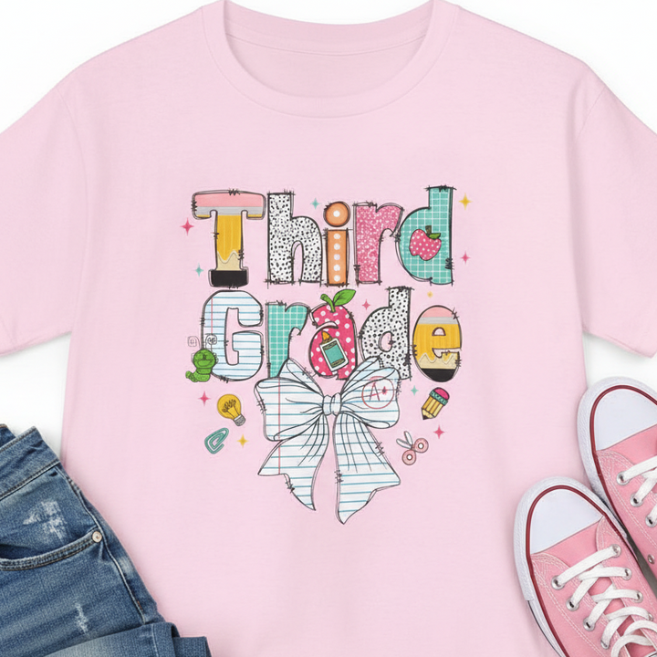A light pink short-sleeve t-shirt with a colorful graphic on the front that reads 'Third Grade' in a playful, hand-drawn style with a striped bow illustration below, shown against a plain white background with jeans and pink converse.