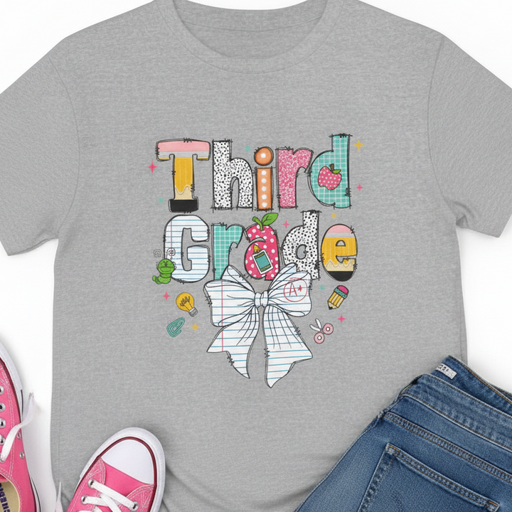 A heather gray short-sleeve T-shirt laid flat with pink sneakers and blue jeans partially visible at the edges. The shirt features a colorful graphic reading 'Third Grade' with school-themed letter designs and a large bow illustration.