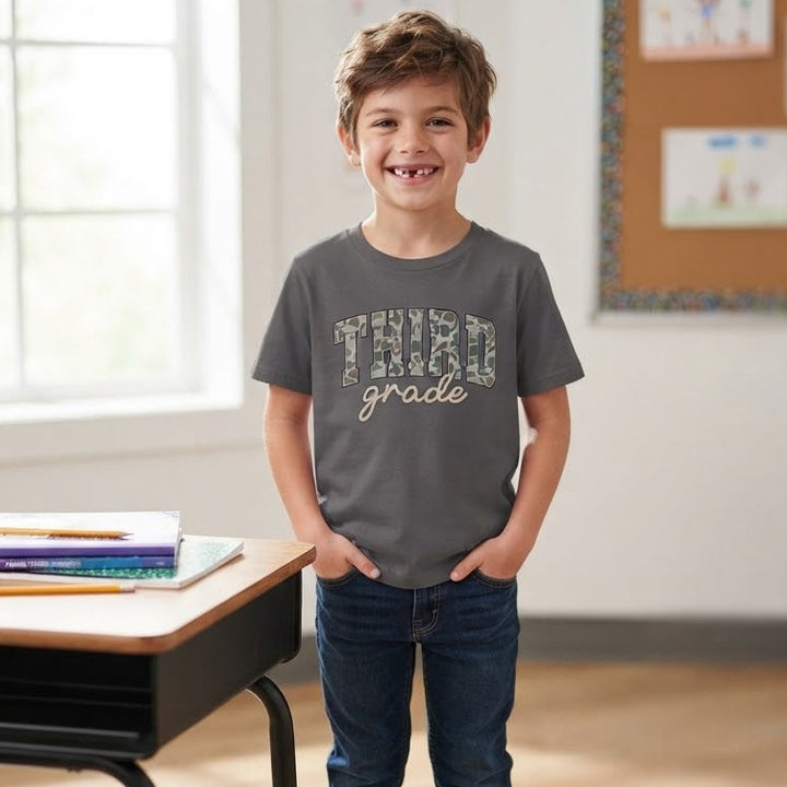 A young boy stands in a bright classroom wearing a dark gray graphic t-shirt that reads 'THIRD grade' in a camouflage pattern, paired with dark blue jeans and gray sneakers.