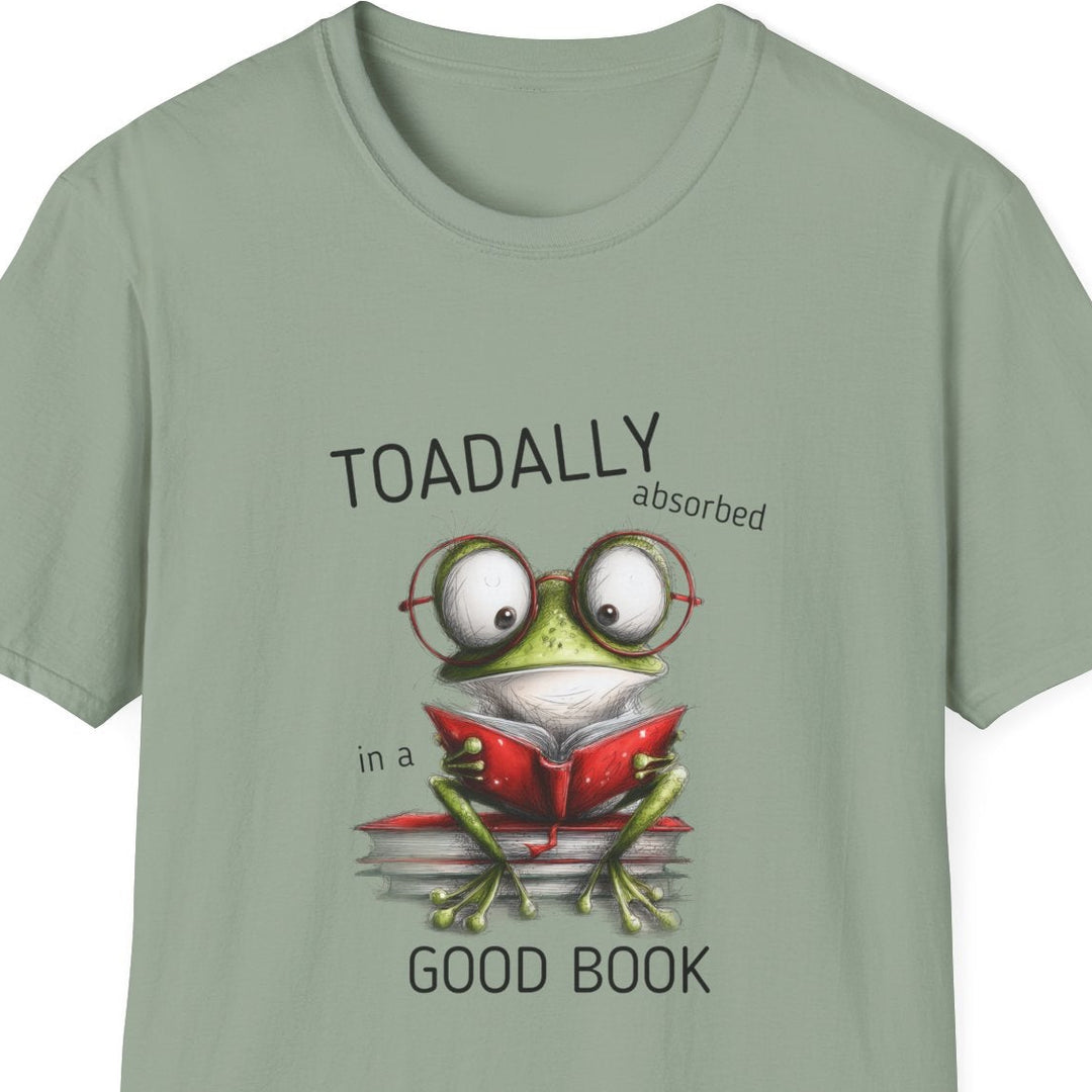 A sage green short-sleeve crewneck t-shirt featuring a cartoon graphic of a green frog wearing round glasses and reading a red book, with the text 'TOADALLY absorbed in a' above and 'GOOD BOOK' below.