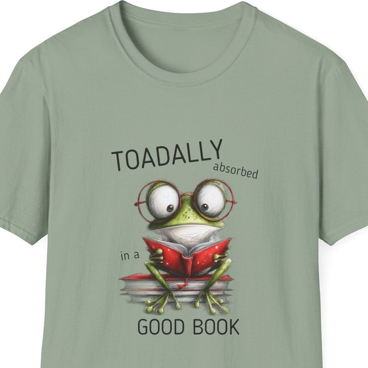 A sage green short-sleeve crewneck t-shirt featuring a cartoon graphic of a green frog wearing round glasses and reading a red book, with the text 'TOADALLY absorbed in a' above and 'GOOD BOOK' below.