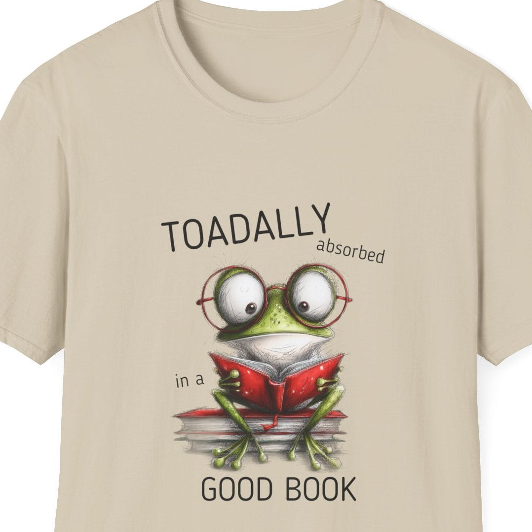 A beige short-sleeve crewneck t-shirt featuring a cartoon graphic of a green frog wearing glasses and reading a red book, with the text 'TOADALLY absorbed in a' and 'GOOD BOOK'.