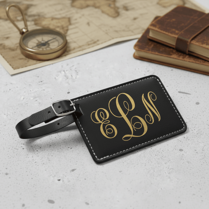 Black luggage tag with gold monogram on a textured surface with books and a compass.