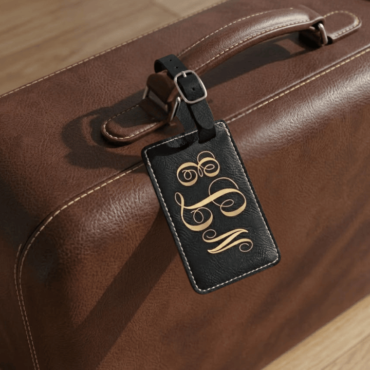 Brown leather suitcase with a black luggage tag featuring gold monogram on a wooden surface.