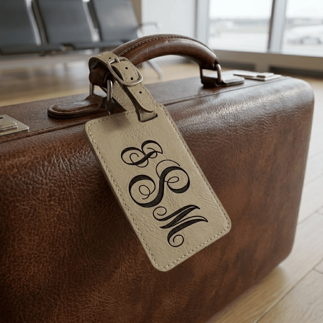 Brown leather suitcase with a beige luggage tag featuring monogram initials in an airport setting.