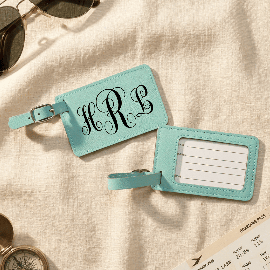 Two turquoise luggage tags with monogram on front and blank label on back sitting on a beige fabric background.