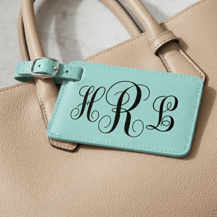 Turquoise luggage tag with 'HRL' monogram engraved in black attached to a beige handbag.
